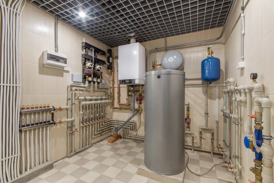 Local technique moderne avec chaudière cylindrique grise, réseau de canalisations blanches avec vannes de régulation, collecteur de distribution, vase d'expansion bleu